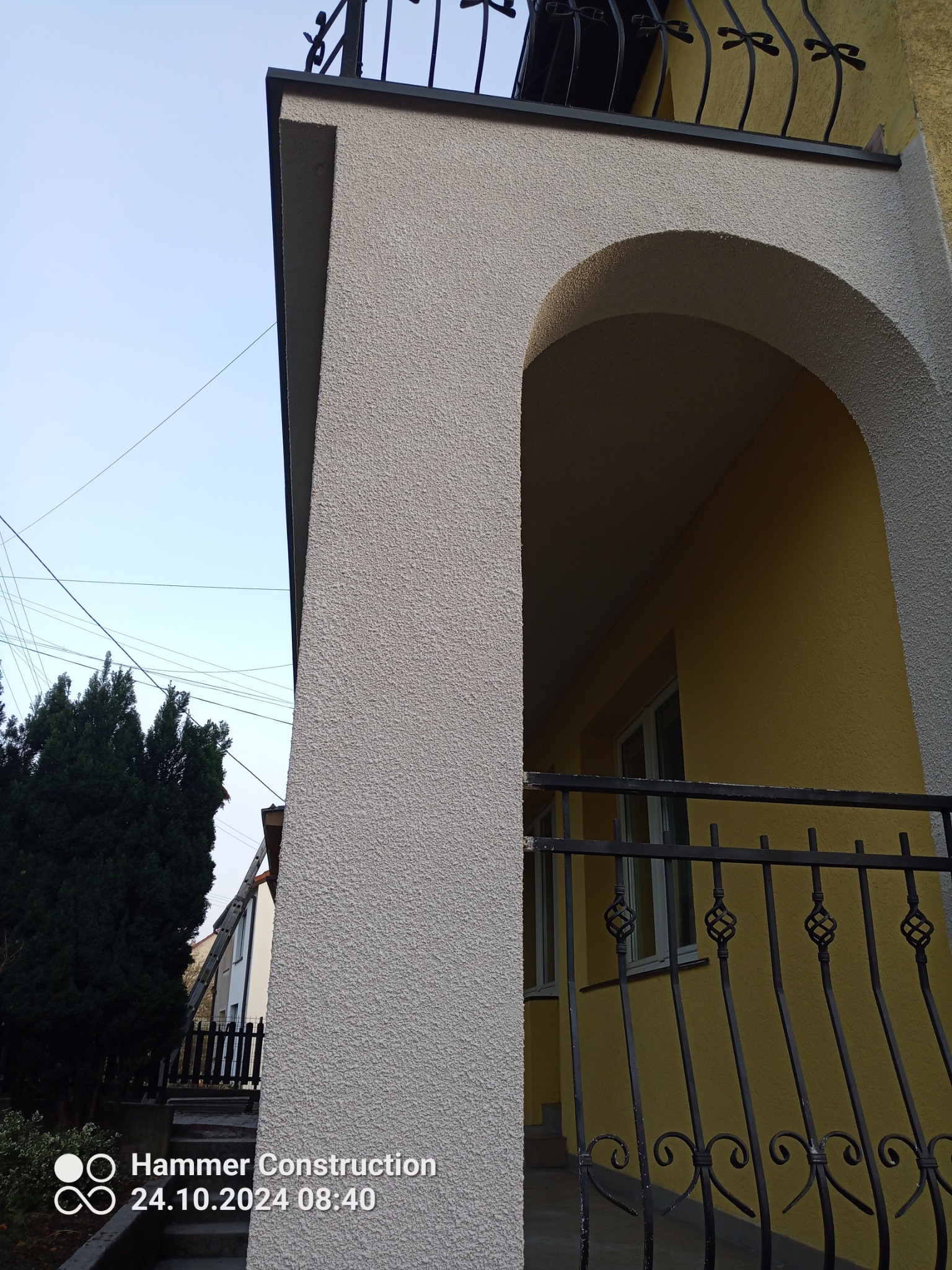 Wykończony balkon z łukowatym wejściem, szara elewacja z fakturą baranka, czarna metalowa balustrada i żółta ściana w tle.