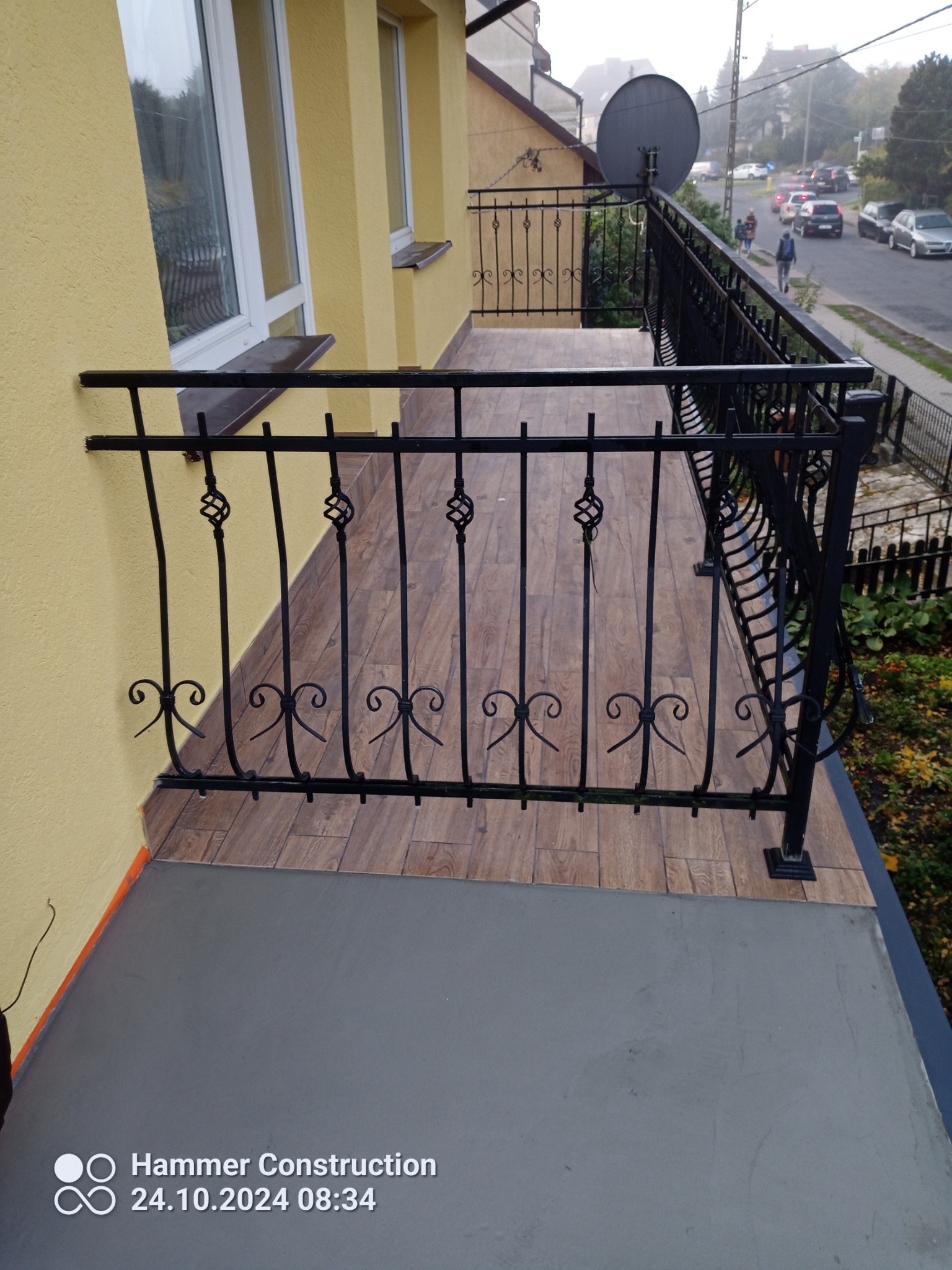 Balkon z czarną, ozdobną balustradą i wykończoną podłogą imitującą drewniane panele, widok na ulicę z zaparkowanymi samochodami i budynkami w tle.