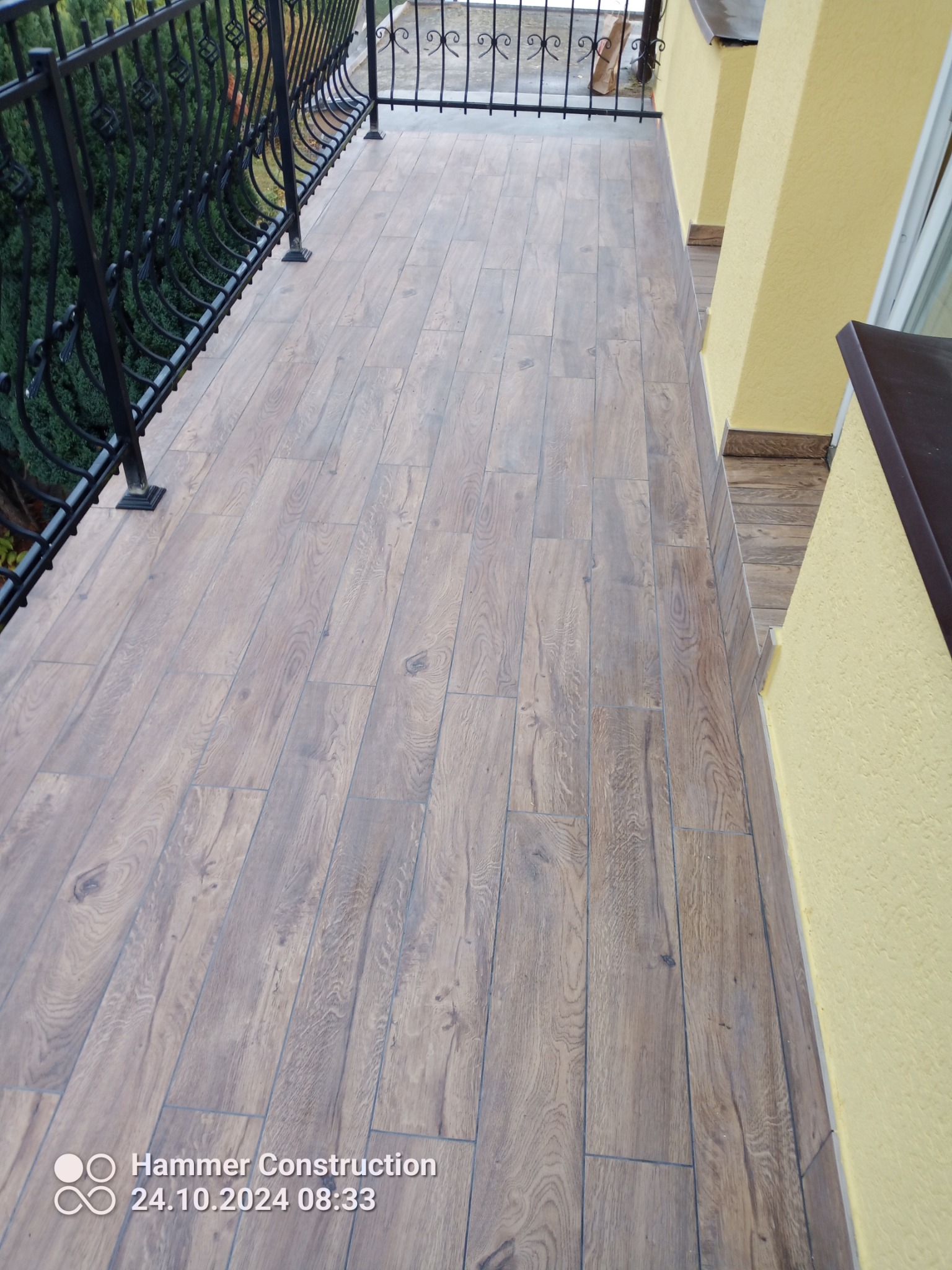 Balkon z podłogą wyłożoną panelami imitującymi drewno, z czarną, ozdobną balustradą i żółtą elewacją budynku.