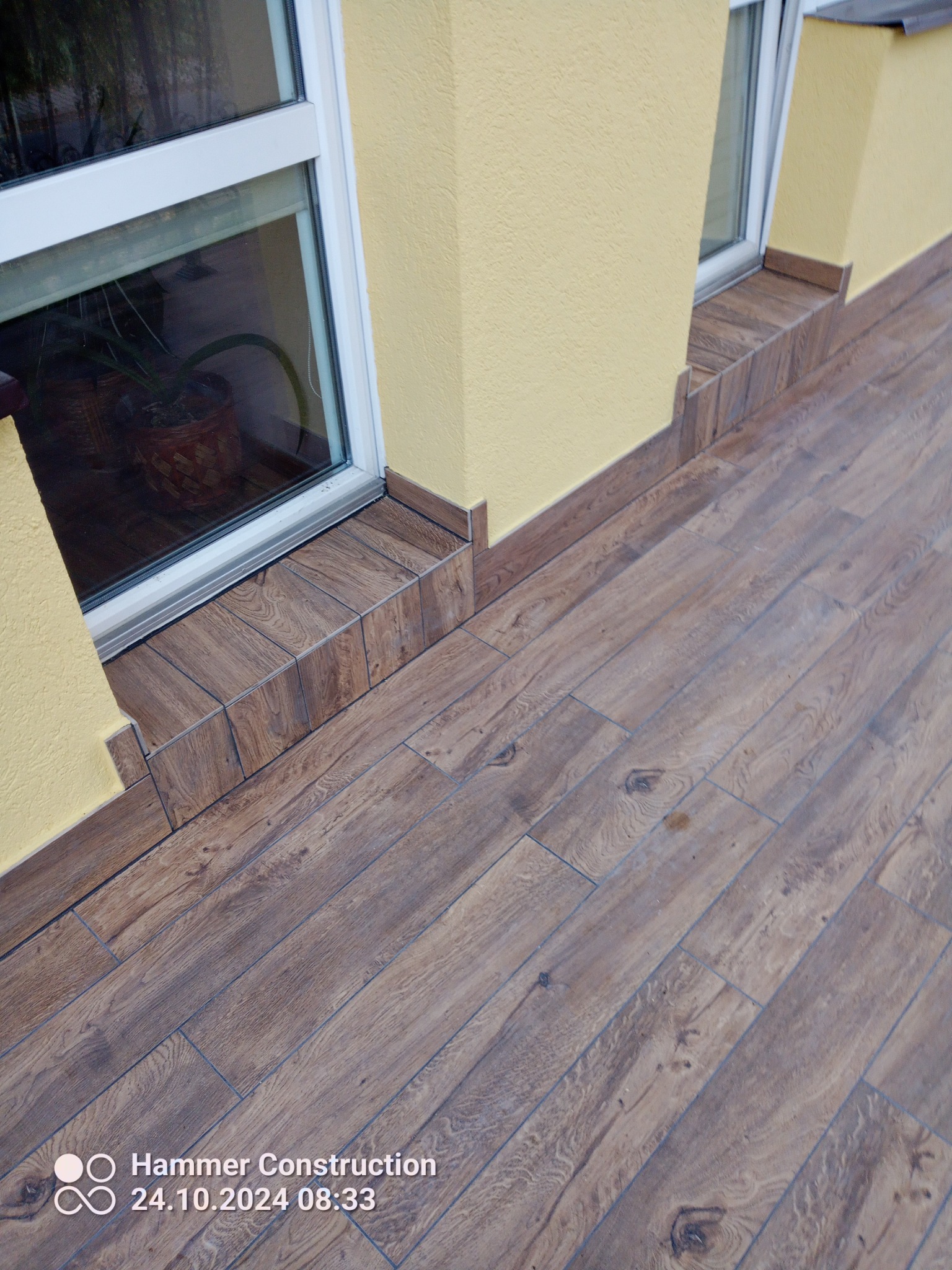 Elewacja budynku z oknem, wykończona płytkami imitującymi drewno na cokole i wokół okna. Żółty tynk strukturalny na ścianie. Widoczne łączenia płytek. Podłoga wyłożona panelami podłogowymi.