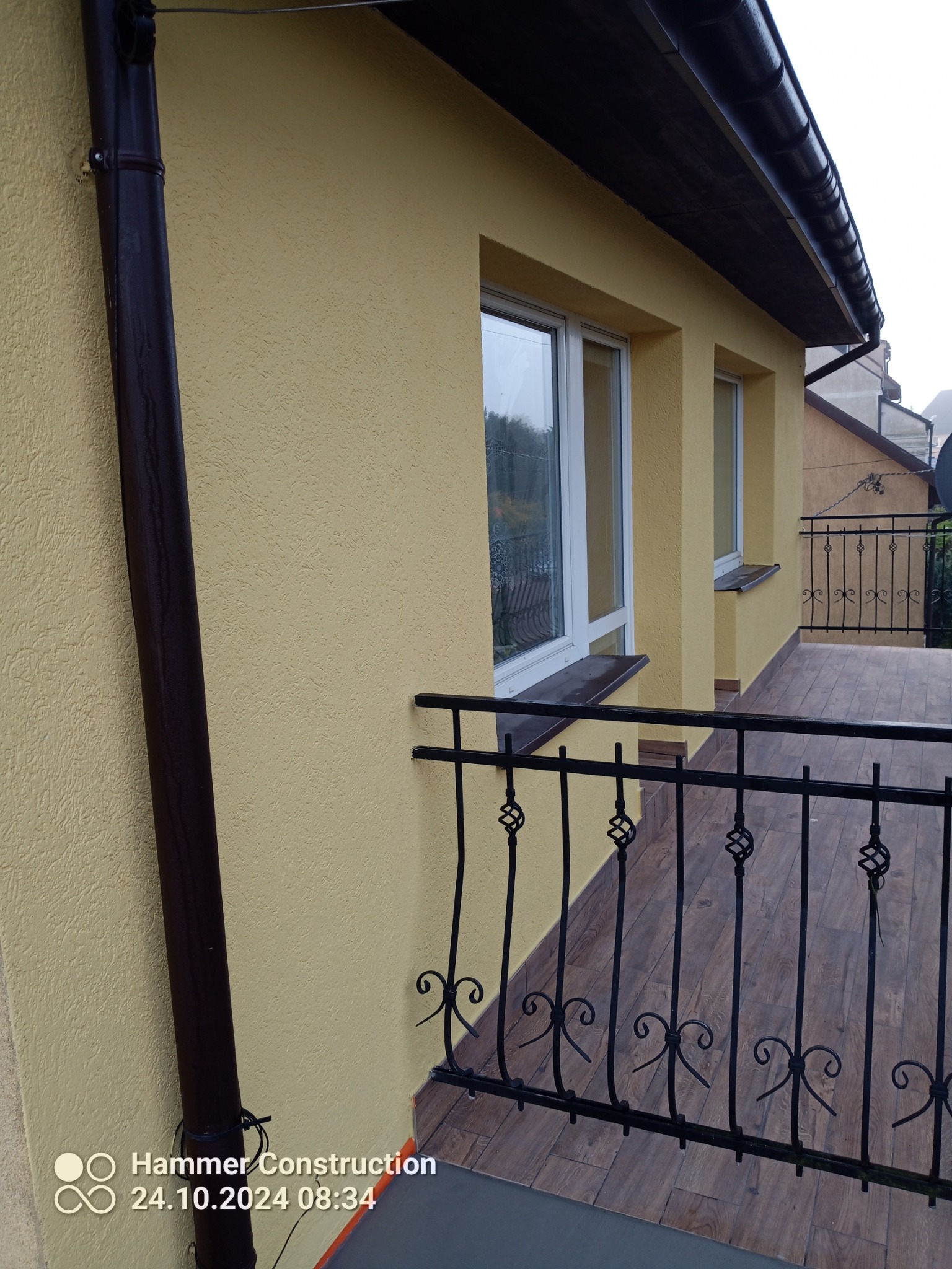 Żółta elewacja budynku z balkonem, brązowa rynna i balustrada balkonu z metalowymi zdobieniami.