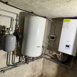 INSTAL-TECH SPÓŁKA Z OGRANICZONĄ ODPOWIEDZIALNOŚCIĄ - Biały zasobnik ciepłej wody użytkowej i piec gazowy Beretta zamontowane w piwnicy z widocznymi rurami instalacji grzewczej i elektrycznej.