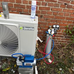 INSTAL-TECH SPÓŁKA Z OGRANICZONĄ ODPOWIEDZIALNOŚCIĄ - Zewnętrzna jednostka pompy ciepła Beretta podłączona do butli z gazem i pompy próżniowej podczas instalacji, widoczny manometr i spray do wykrywania nieszczelności, tło to ceglany mur i ziemia...
