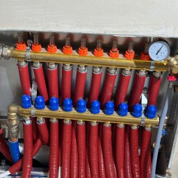 INSTAL-TECH SPÓŁKA Z OGRANICZONĄ ODPOWIEDZIALNOŚCIĄ - Rozdzielacz ogrzewania podłogowego z rurami w czerwonej izolacji i niebieskimi zaworami, widoczny manometr i odpowietrzniki na tle surowej ściany.