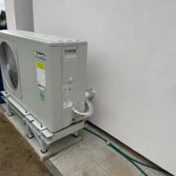 Zewnętrzna jednostka pompy ciepła Beretta zamontowana na metalowej konstrukcji przy ścianie budynku. Widoczne rury w izolacji aluminiowej oraz zielony wąż ogrodowy. W tle pojemniki na śmieci.