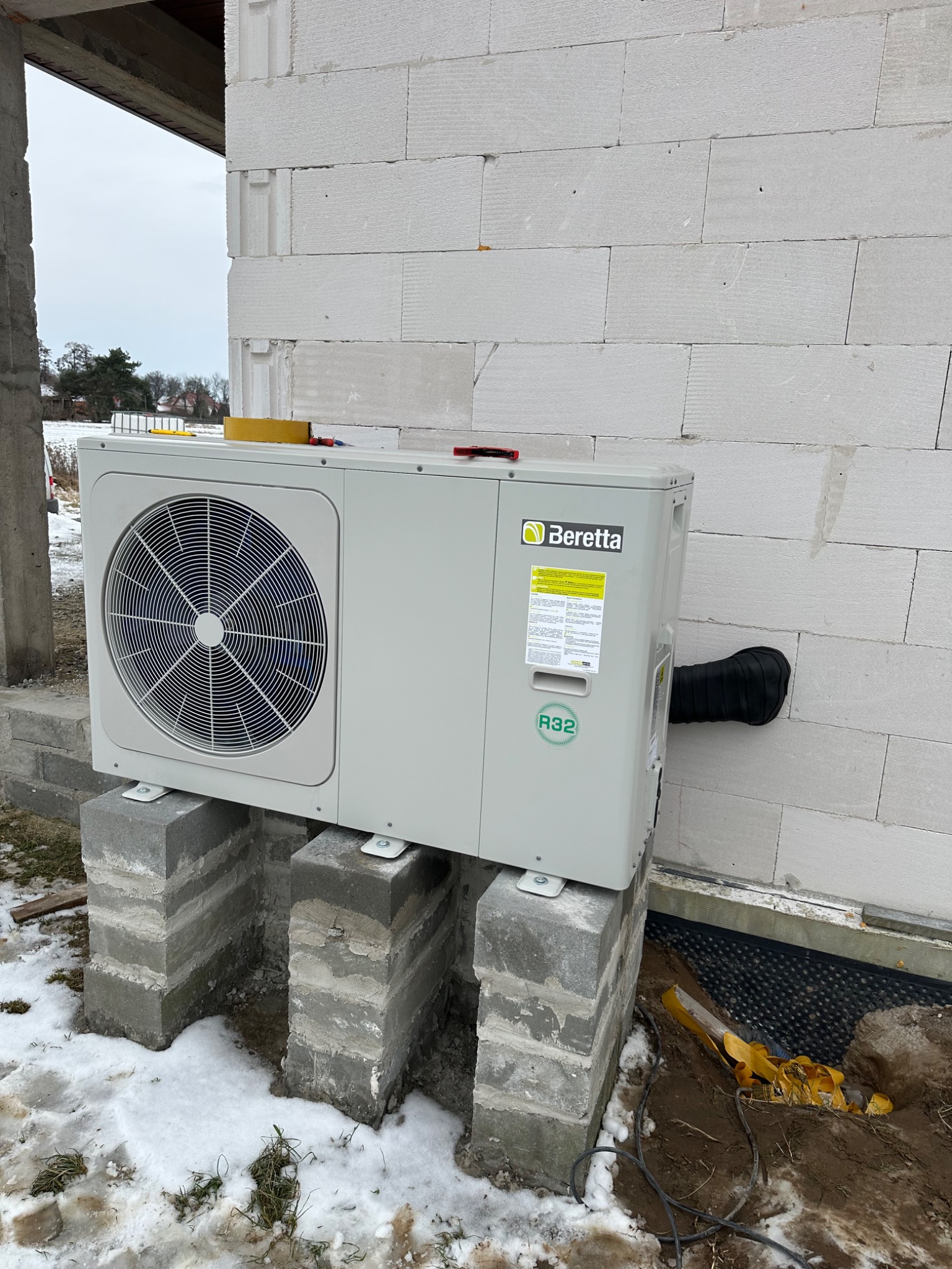 Zewnętrzna jednostka klimatyzacyjna Beretta, zamontowana na betonowych blokach przed surową ścianą budynku z betonu komórkowego, widoczne elementy montażowe i czarny przewód odprowadzający.