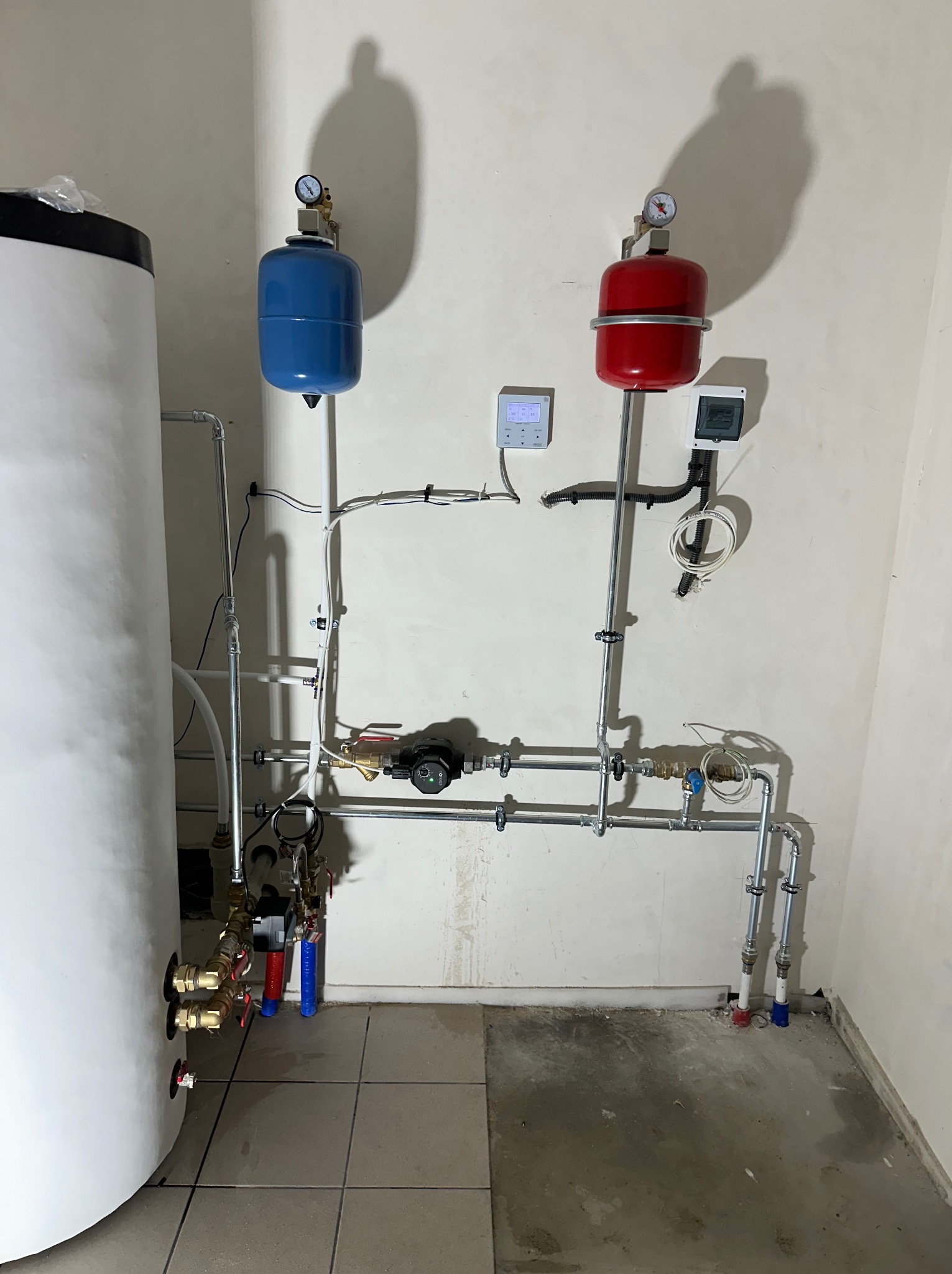 Instalacja grzewcza w kotłowni: biały zasobnik ciepłej wody, czerwony i niebieski zbiornik przeponowy, rury z zaworami, manometry oraz sterowniki na ścianie.