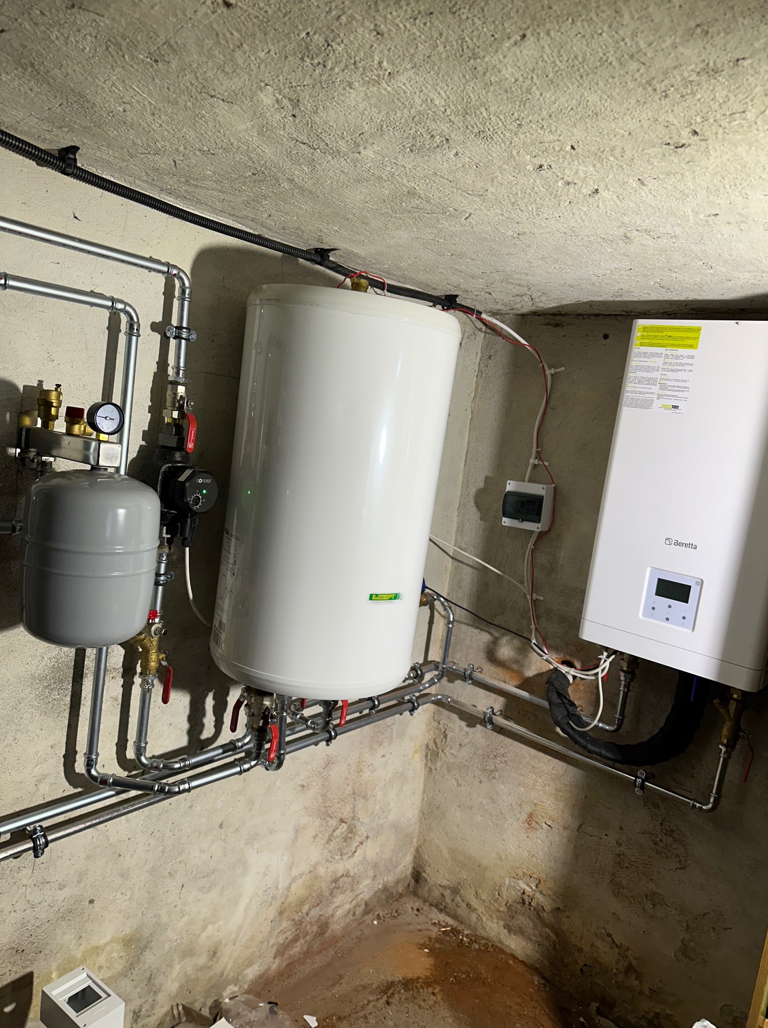 Biały zasobnik ciepłej wody użytkowej i piec gazowy Beretta zamontowane w piwnicy z widocznymi rurami instalacji grzewczej i elektrycznej.