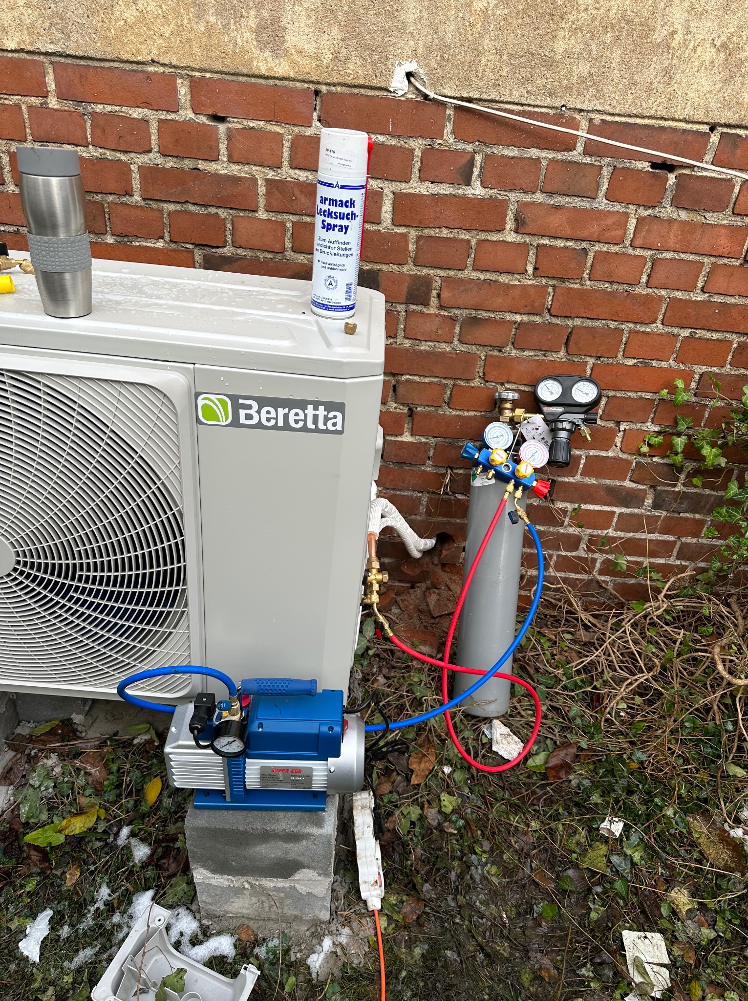 Zewnętrzna jednostka pompy ciepła Beretta podłączona do butli z gazem i pompy próżniowej podczas instalacji, widoczny manometr i spray do wykrywania nieszczelności, tło to ceglany mur i ziemia...