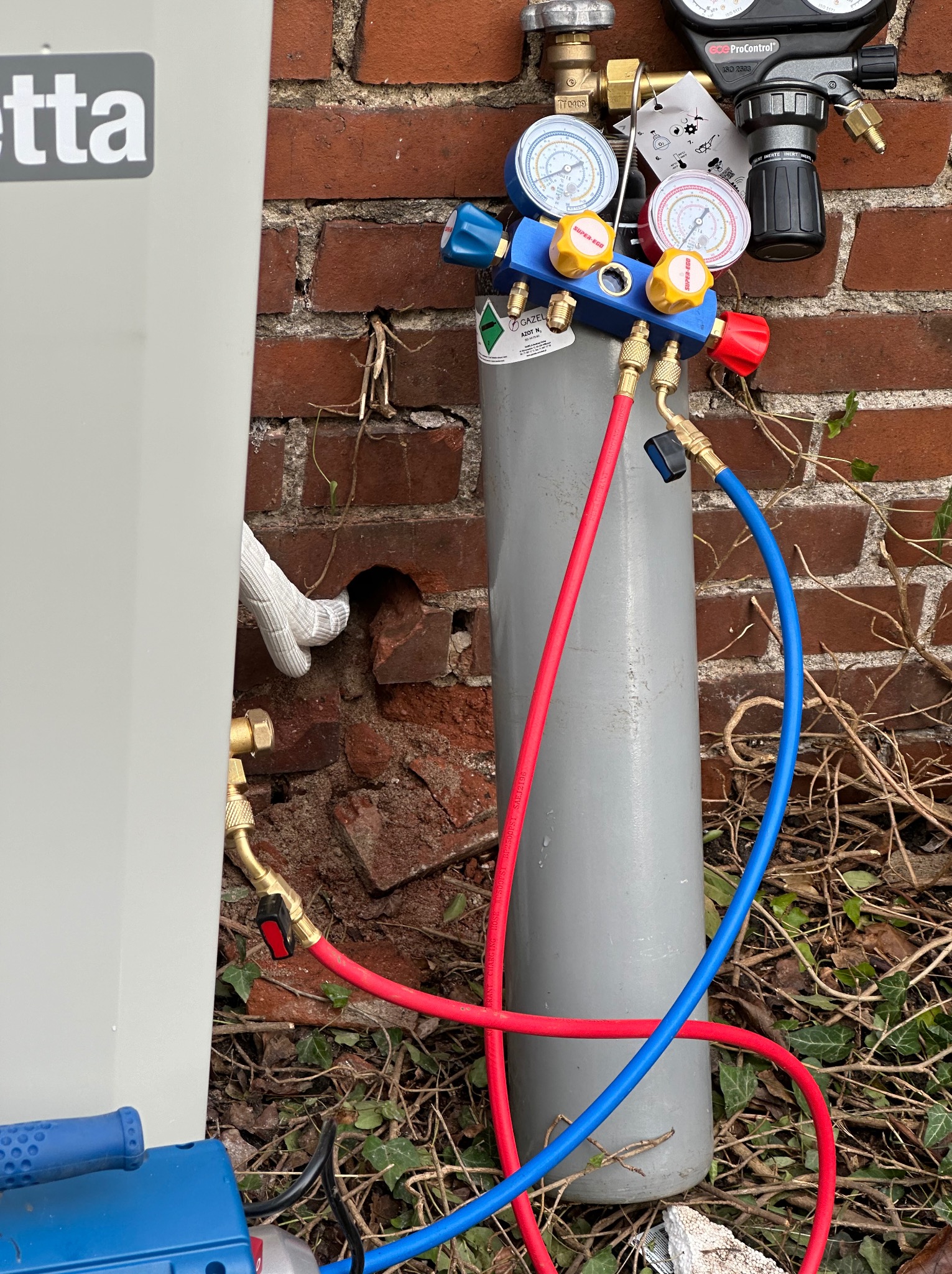 Podłączenie butli z gazem technicznym do pompy ciepła, widoczne manometry, węże w kolorze czerwonym i niebieskim, ceglana ściana w tle, instalacja na zewnątrz budynku, przyłącze gazowe.
