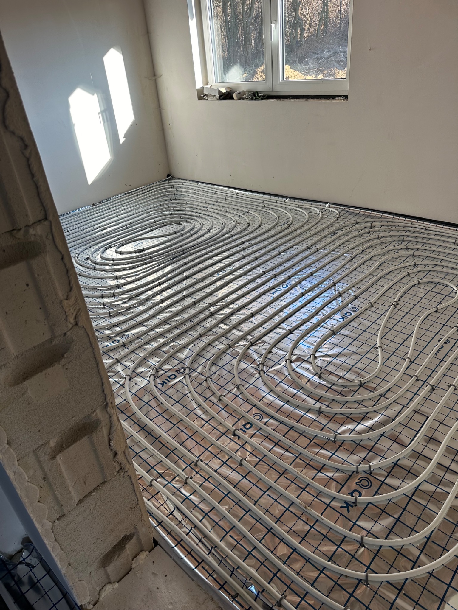 Białe rury ogrzewania podłogowego ułożone spiralnie na folii z nadrukowaną siatką, widoczne przez okno światło słoneczne i surowy mur w tle.