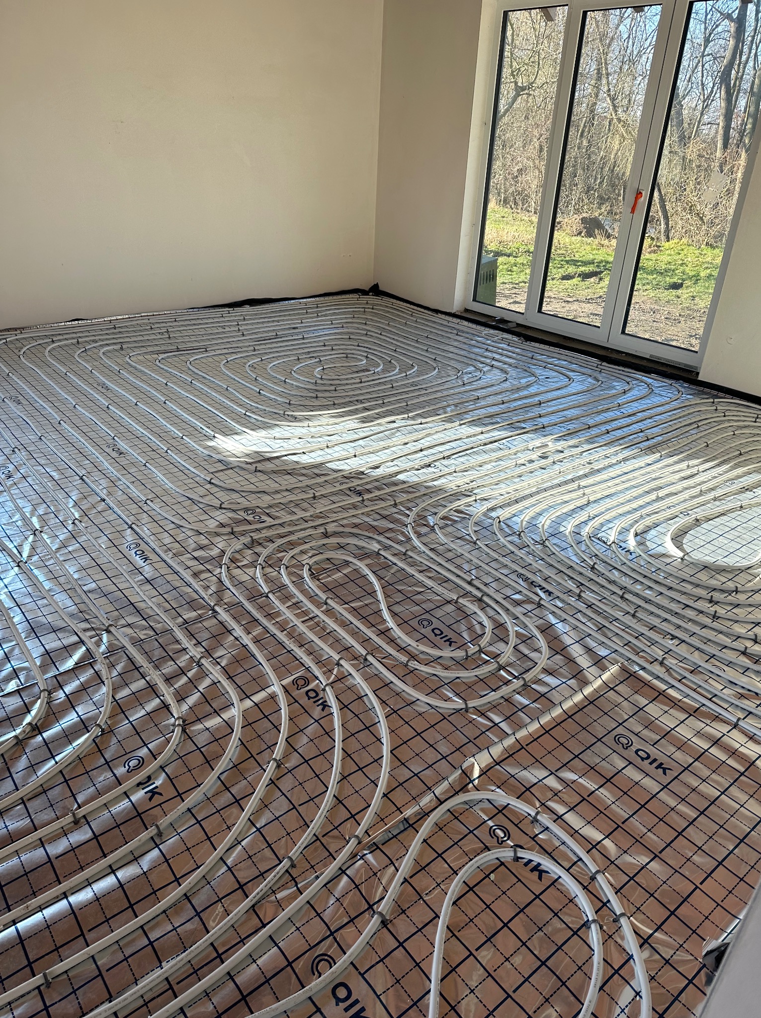 Białe rurki ogrzewania podłogowego ułożone spiralnie na folii z nadrukiem kratki w jasnym, pustym pomieszczeniu z dużym oknem balkonowym.