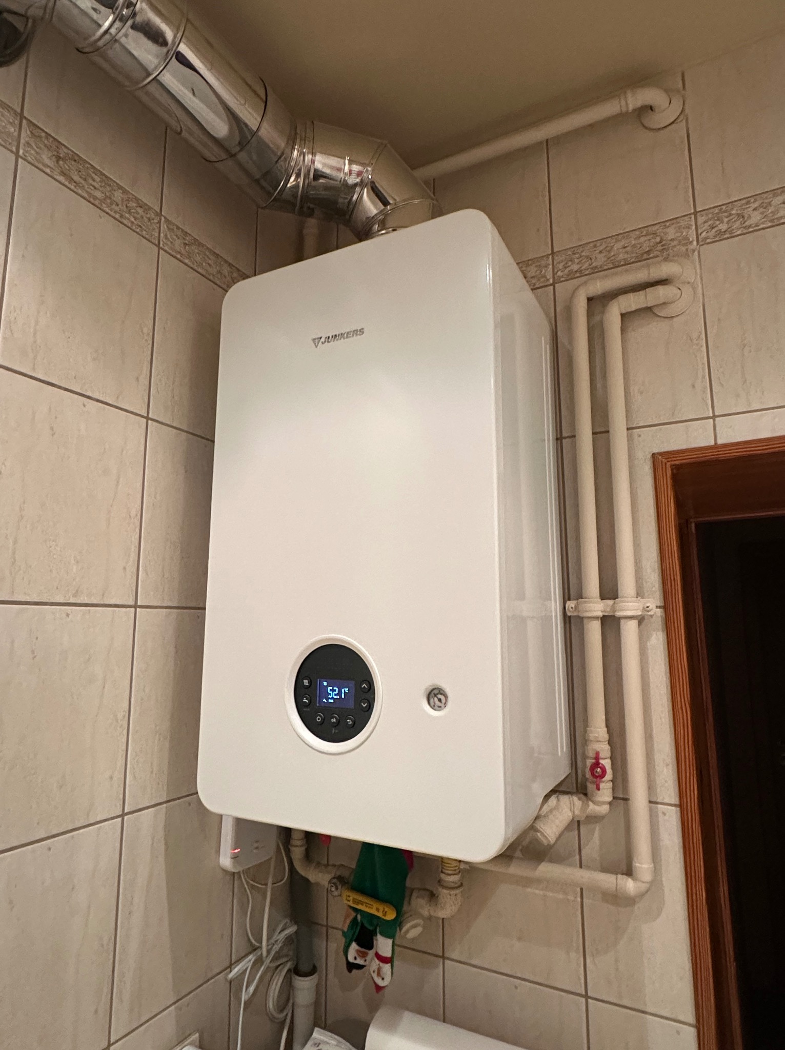 Biały piec gazowy Junkers zamontowany na kafelkowanej ścianie, widoczne rury odprowadzające spaliny i instalacja hydrauliczna z zaworem z czerwoną rączką, na wyświetlaczu pieca widoczna temperatura...