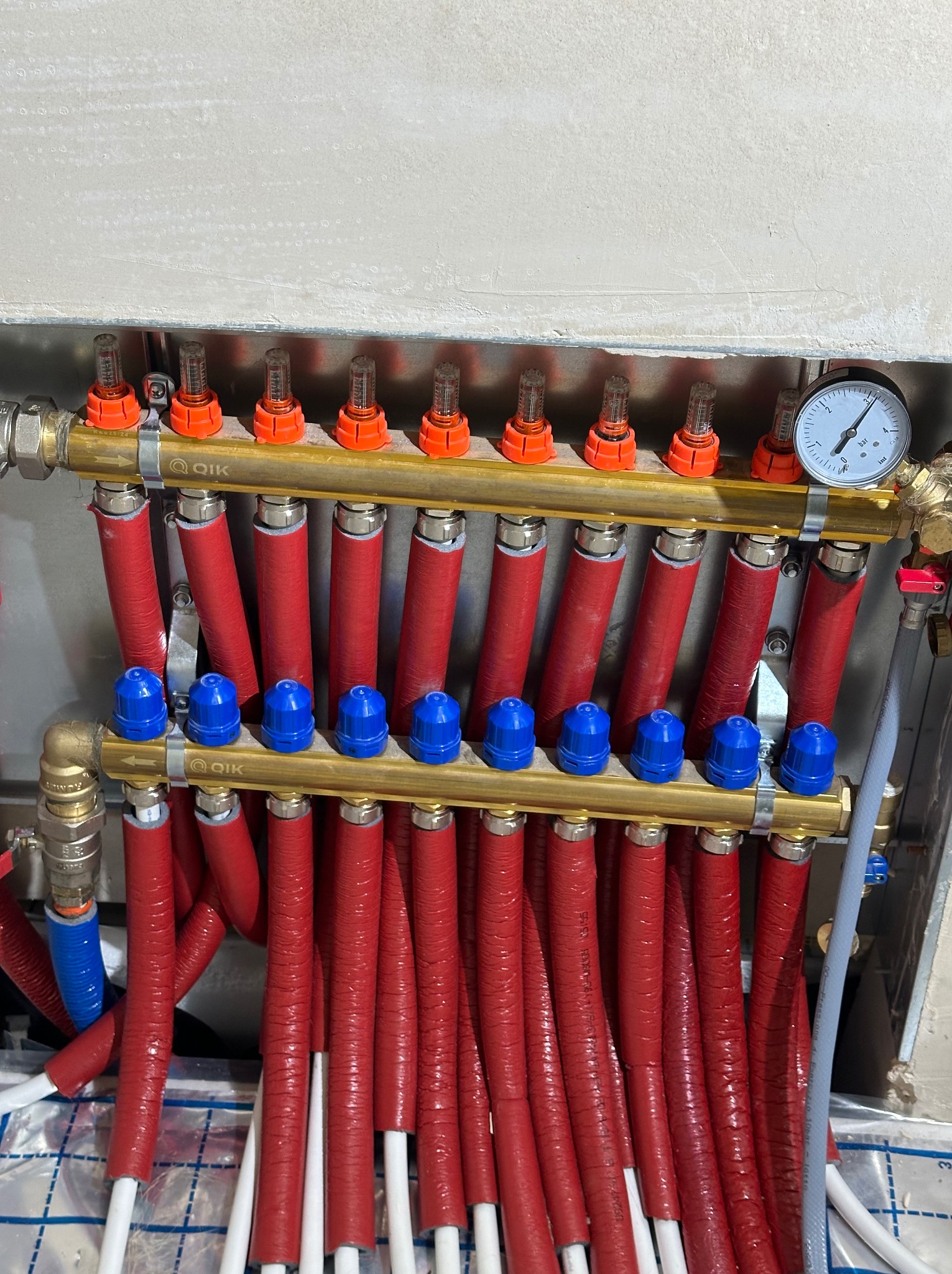 Rozdzielacz ogrzewania podłogowego z rurami w czerwonej izolacji i niebieskimi zaworami, widoczny manometr i odpowietrzniki na tle surowej ściany.