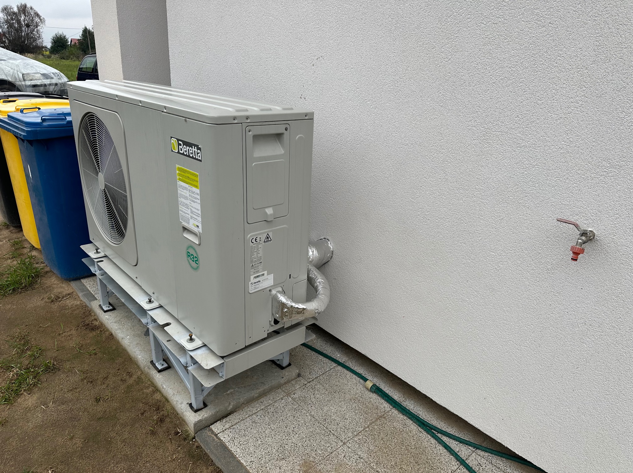 Zewnętrzna jednostka pompy ciepła Beretta zamontowana na metalowej konstrukcji przy ścianie budynku. Widoczne rury w izolacji aluminiowej oraz zielony wąż ogrodowy. W tle pojemniki na śmieci.