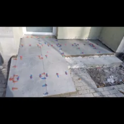 Układanie szarych płytek na zewnątrz budynku, widoczne krzyżyki dystansowe w kolorze niebieskim i pomarańczowym, fragment kostki brukowej obok.