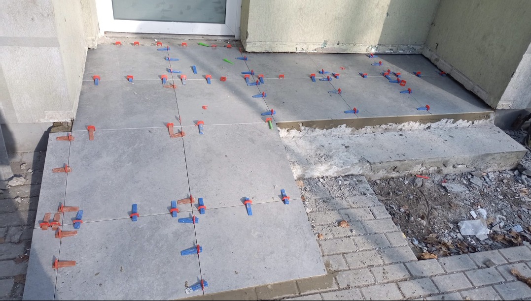 Kładzenie szarych płytek na zewnątrz budynku, widoczne krzyżyki dystansowe i poziomica sznurkowa, fragment chodnika z kostki brukowej obok