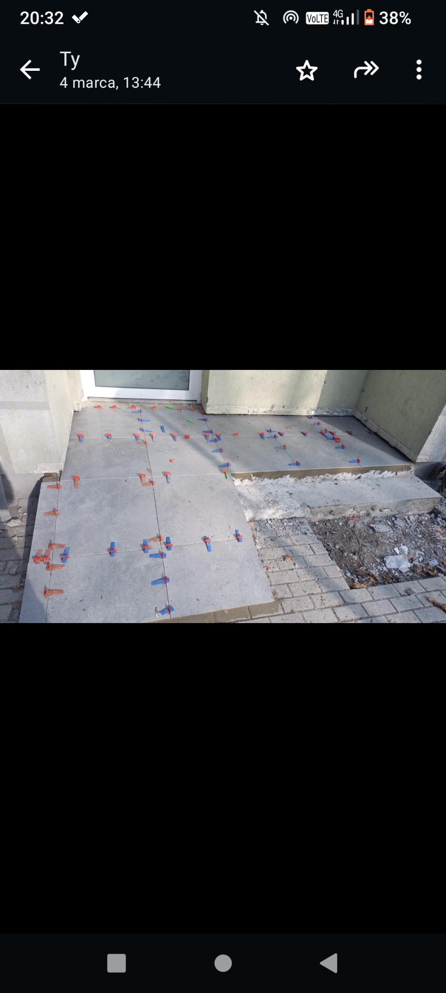 Układanie szarych płytek na zewnątrz budynku, widoczne krzyżyki dystansowe w kolorze niebieskim i pomarańczowym, fragment kostki brukowej obok.