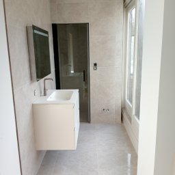 Fachmar - Jasna łazienka z beżowymi płytkami na ścianach i podłodze, biała szafka z umywalką i lustrem, kabina prysznicowa z czarnymi ramami i okno z białymi ramami.