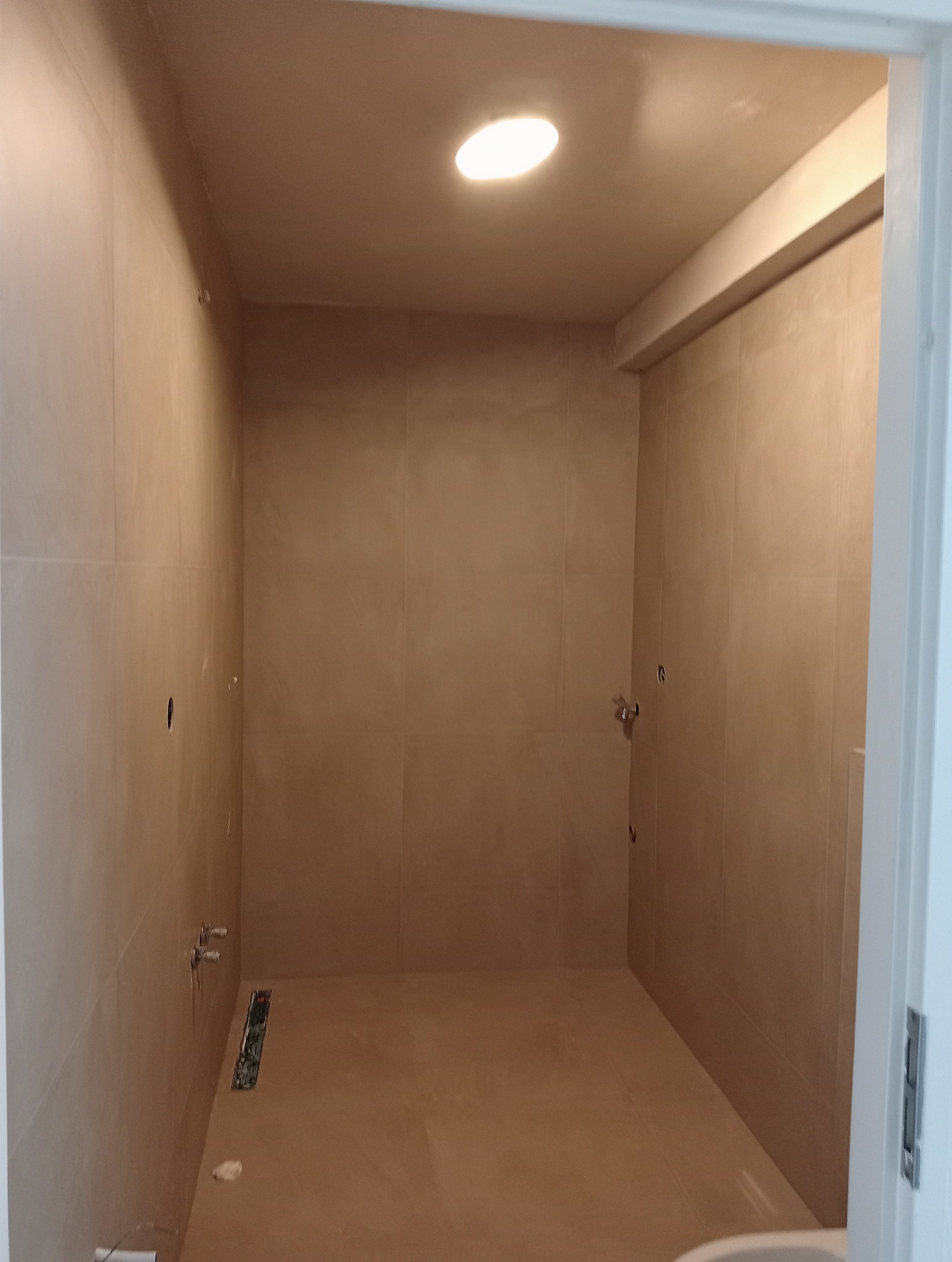 Wykończona łazienka z beżowymi płytkami na ścianach i podłodze, widoczny odpływ liniowy, przygotowane punkty pod armaturę, oświetlenie sufitowe.