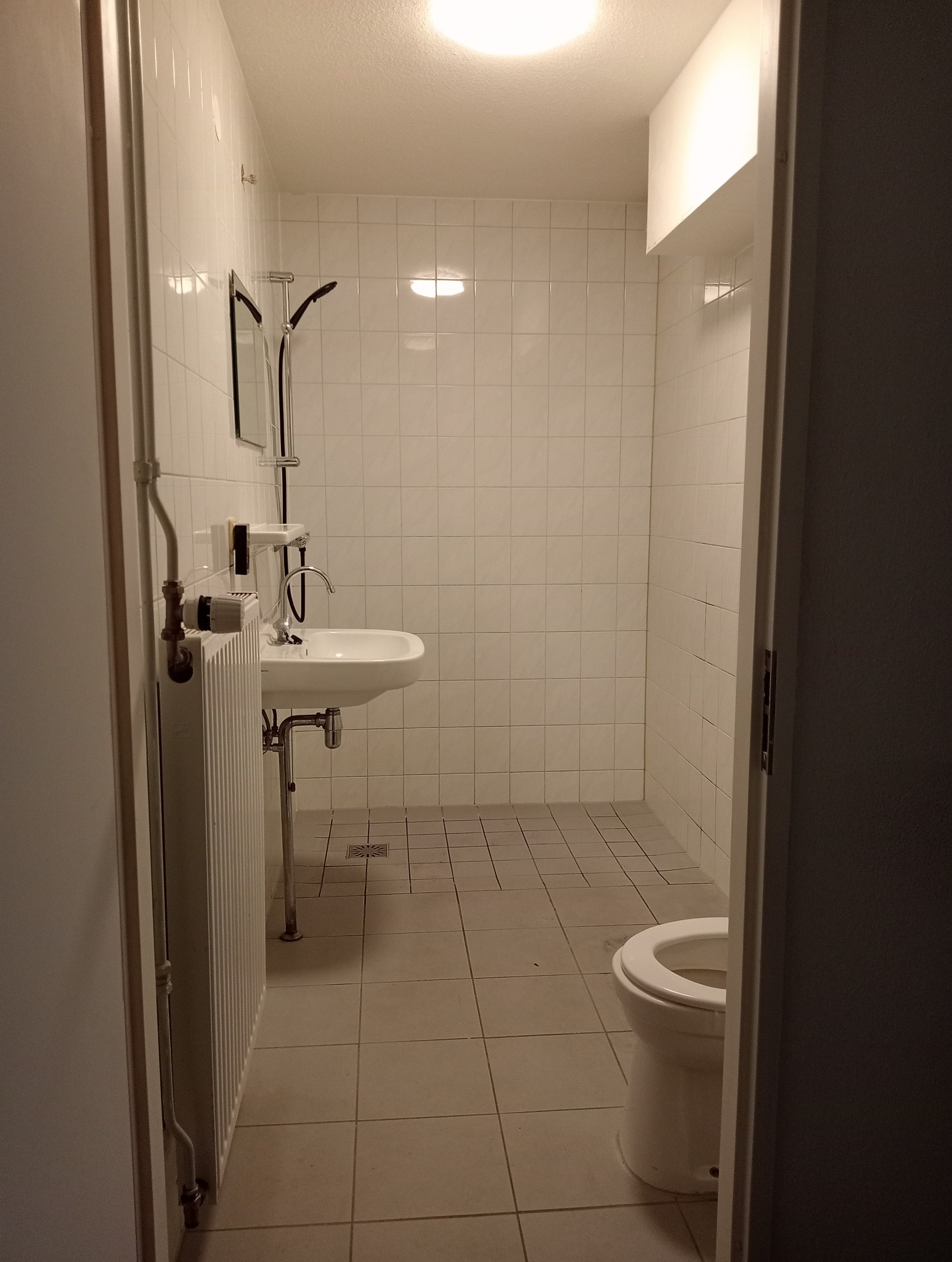 Wnętrze łazienki z białymi kwadratowymi płytkami na ścianach i podłodze, widoczna umywalka, grzejnik żeberkowy i toaleta w rogu, oświetlenie sufitowe.