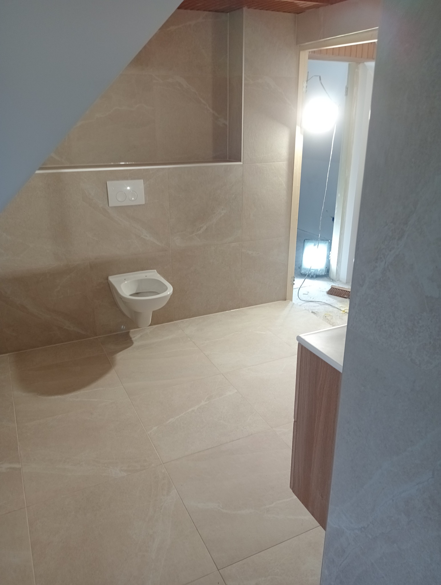 Beżowe płytki podłogowe i ścienne w łazience z podwieszaną toaletą, widok na otwarte drzwi z oświetleniem budowlanym w tle.