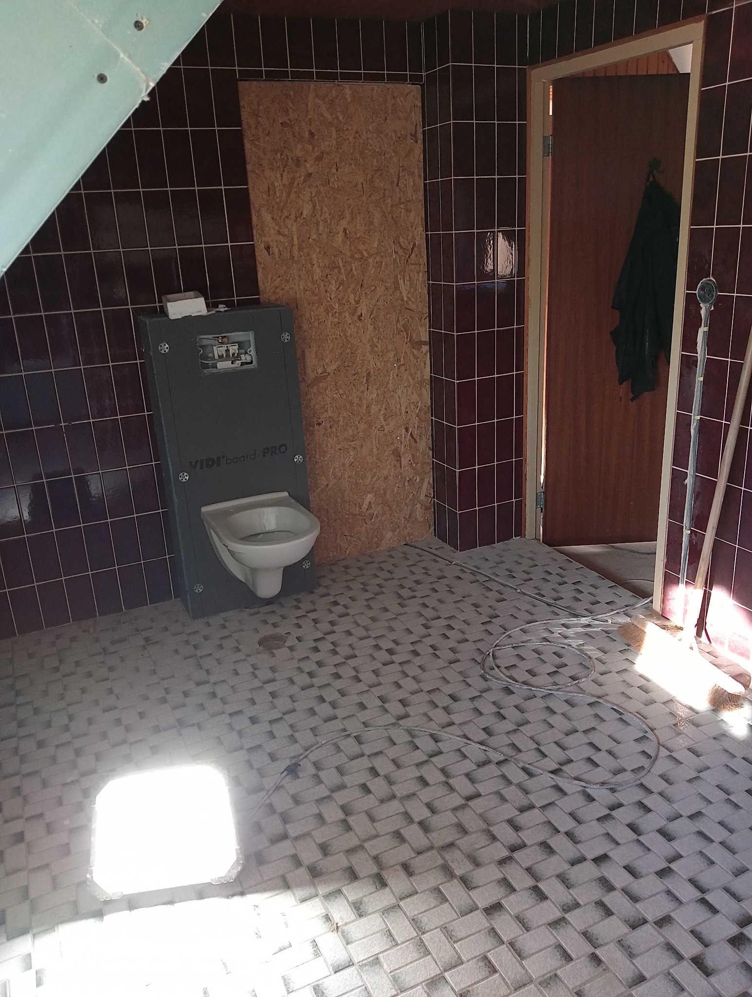 Wnętrze łazienki w trakcie remontu, widoczne ciemne płytki na ścianach, jasne płytki na podłodze ułożone w wzór, zamontowany stelaż podtynkowy z częściowo zamontowaną toaletą, płyta OSB na ścianie,...