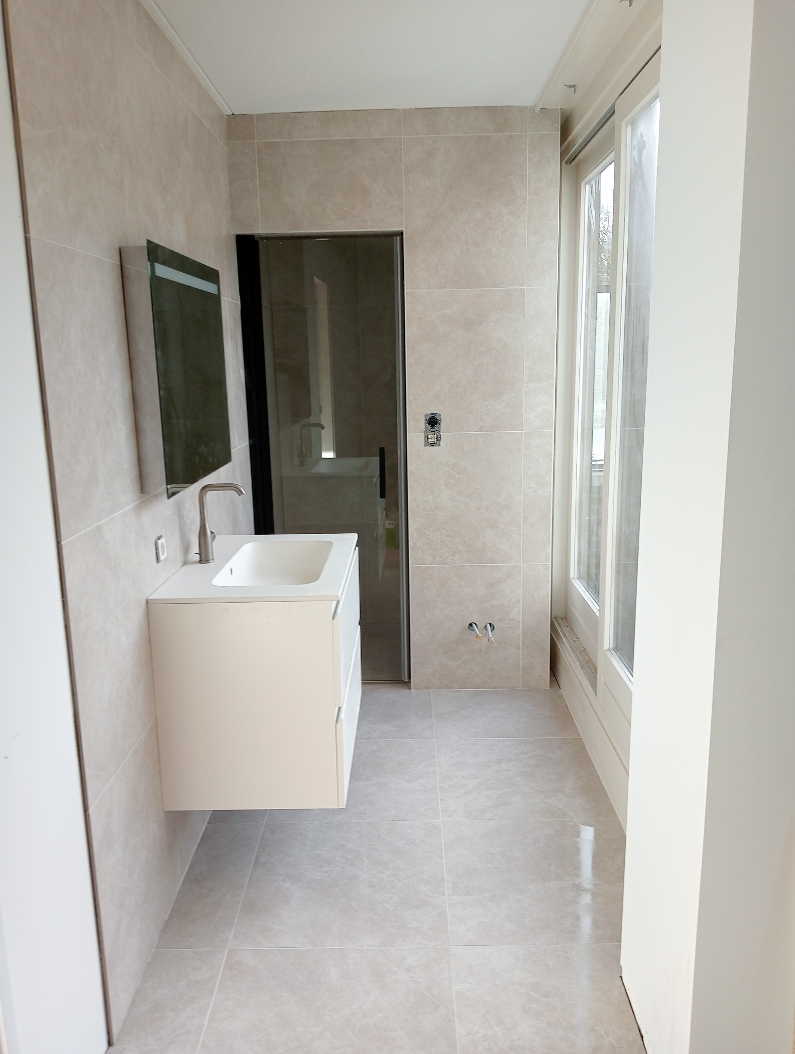 Jasna łazienka z beżowymi płytkami na ścianach i podłodze, biała szafka z umywalką i lustrem, kabina prysznicowa z czarnymi ramami i okno z białymi ramami.