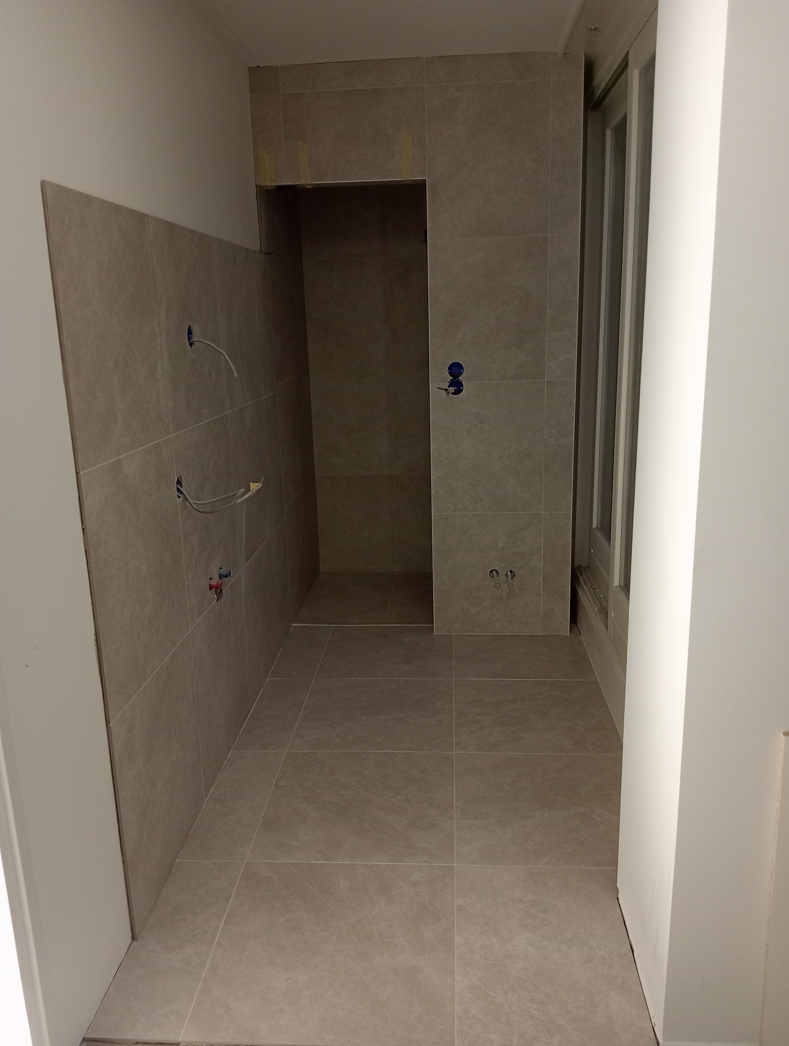 Łazienka w trakcie remontu z beżowymi płytkami ceramicznymi na ścianach i podłodze, widoczne wyprowadzenia instalacji wodnej i elektrycznej, oraz wnęka prysznicowa.