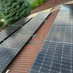 Panele fotowoltaiczne zamontowane na pofałdowanym, rdzawym dachu z widokiem na zalesione wzgórze w tle.