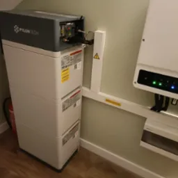 Trzy moduły magazynu energii Pylontech ustawione jeden na drugim, z widocznymi kablami i naklejkami ostrzegawczymi, obok falownik fotowoltaiczny na ścianie w pomieszczeniu z drewnianą podłogą.