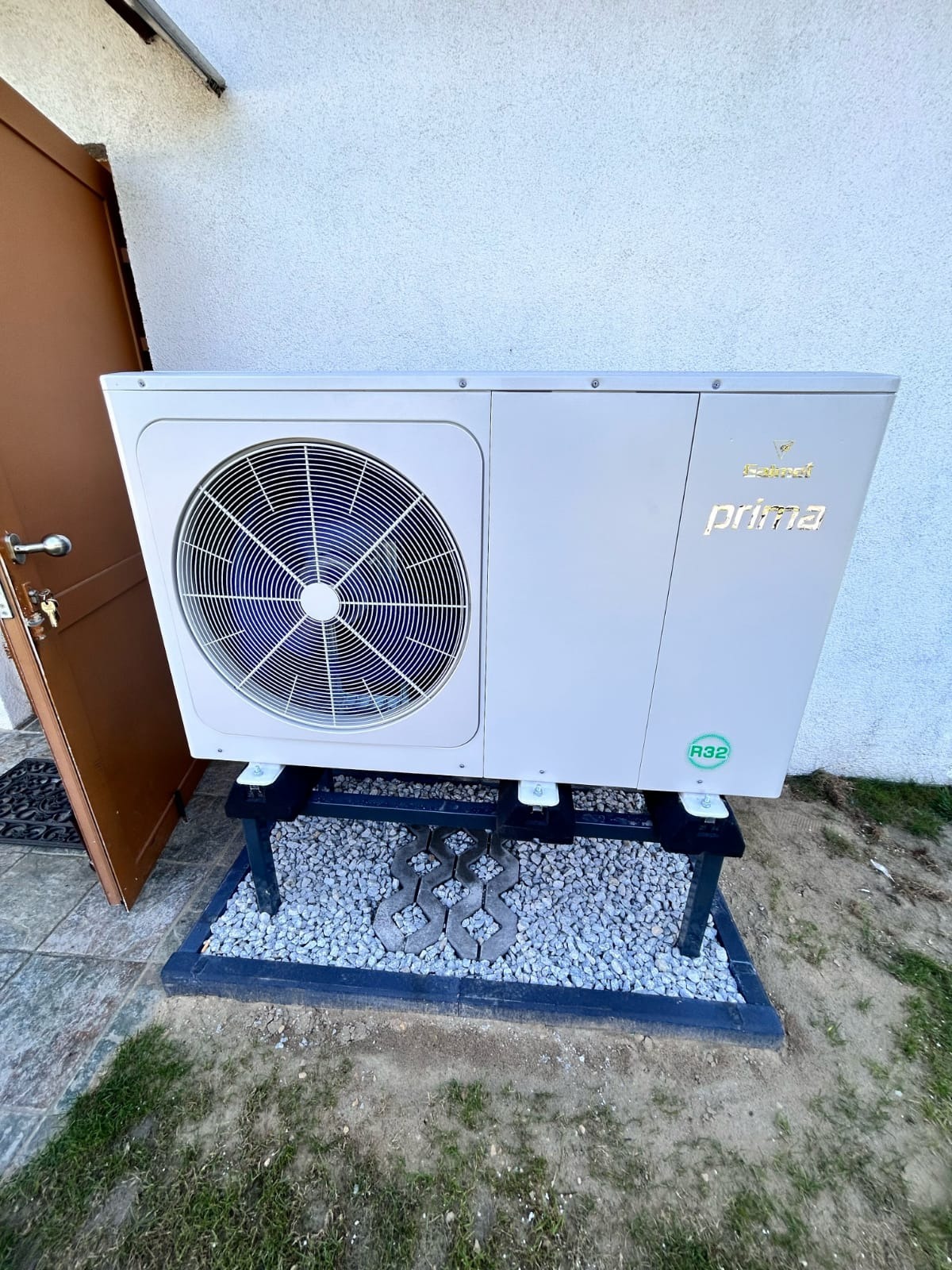 Biała pompa ciepła marki Prima zamontowana na czarnym stelażu z szarym żwirem pod spodem, stojąca przed białą ścianą budynku obok brązowych drzwi.
