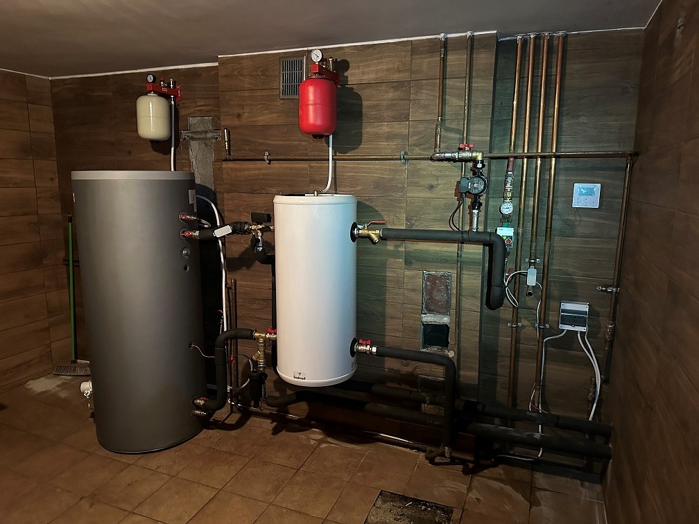 Instalacja grzewcza w kotłowni z dwoma zbiornikami akumulacyjnymi: szarym i białym, licznymi rurami miedzianymi i czarnymi, manometrami i osprzętem na tle ściany wyłożonej brązowymi płytkami...