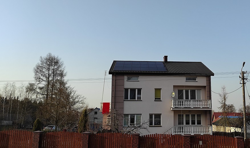 Dwupiętrowy dom z balkonami i panelami słonecznymi na dachu, widoczny zza drewnianego płotu w słoneczny dzień.