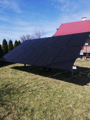 Czarny panel fotowoltaiczny zamontowany na metalowej konstrukcji na trawniku przed domem z czerwonym dachem, widoczne drzewa w tle, słoneczny dzień.