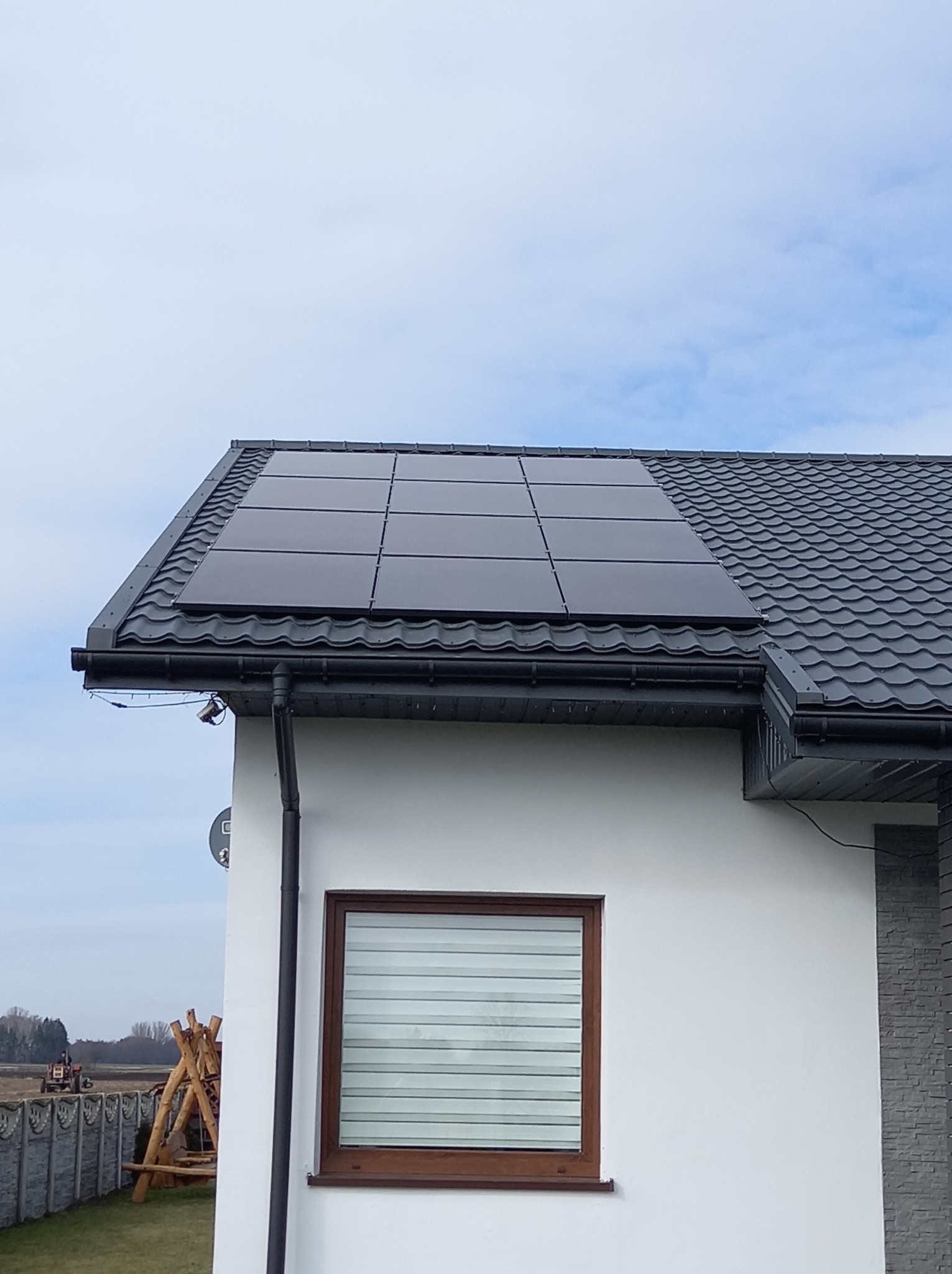 Panele fotowoltaiczne na dachu domu z szarej blachodachówki, widoczne okno z brązową ramą i fragment elewacji wykończonej imitacją kamienia, w tle pole i drzewa.