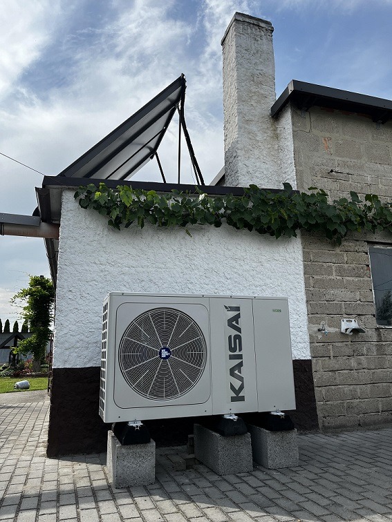 Zewnętrzna jednostka pompy ciepła marki Kaisai, umieszczona na betonowych blokach przy ścianie budynku z fakturą baranka, z widocznym panelem solarnym na dachu i porośniętym pnączem fragmentem...