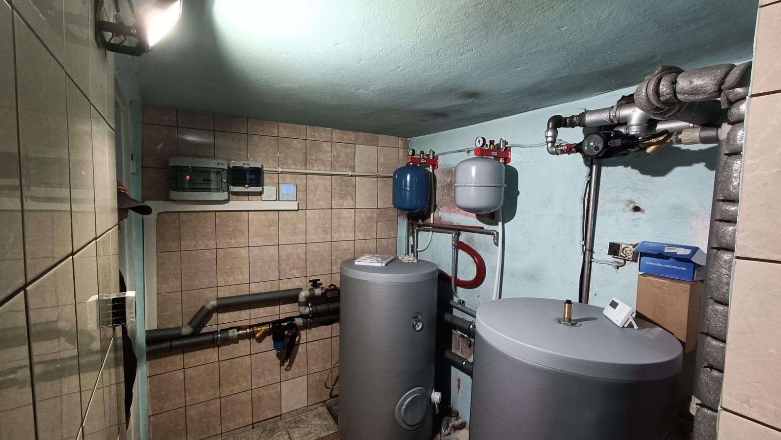 Instalacja dwóch szarych zbiorników akumulacyjnych ciepła z niebieskim i szarym naczyniem przeponowym, rurami, zaworami i sterownikami w pomieszczeniu z beżowymi płytkami i oświetleniem sufitowym.