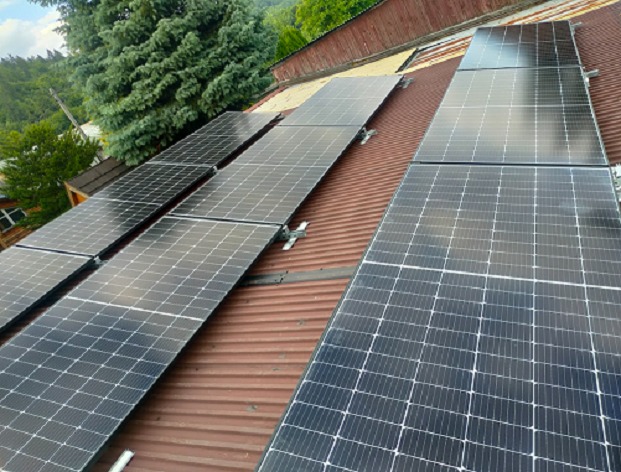 Panele fotowoltaiczne zamontowane na pofałdowanym, rdzawym dachu z widokiem na zalesione wzgórze w tle.