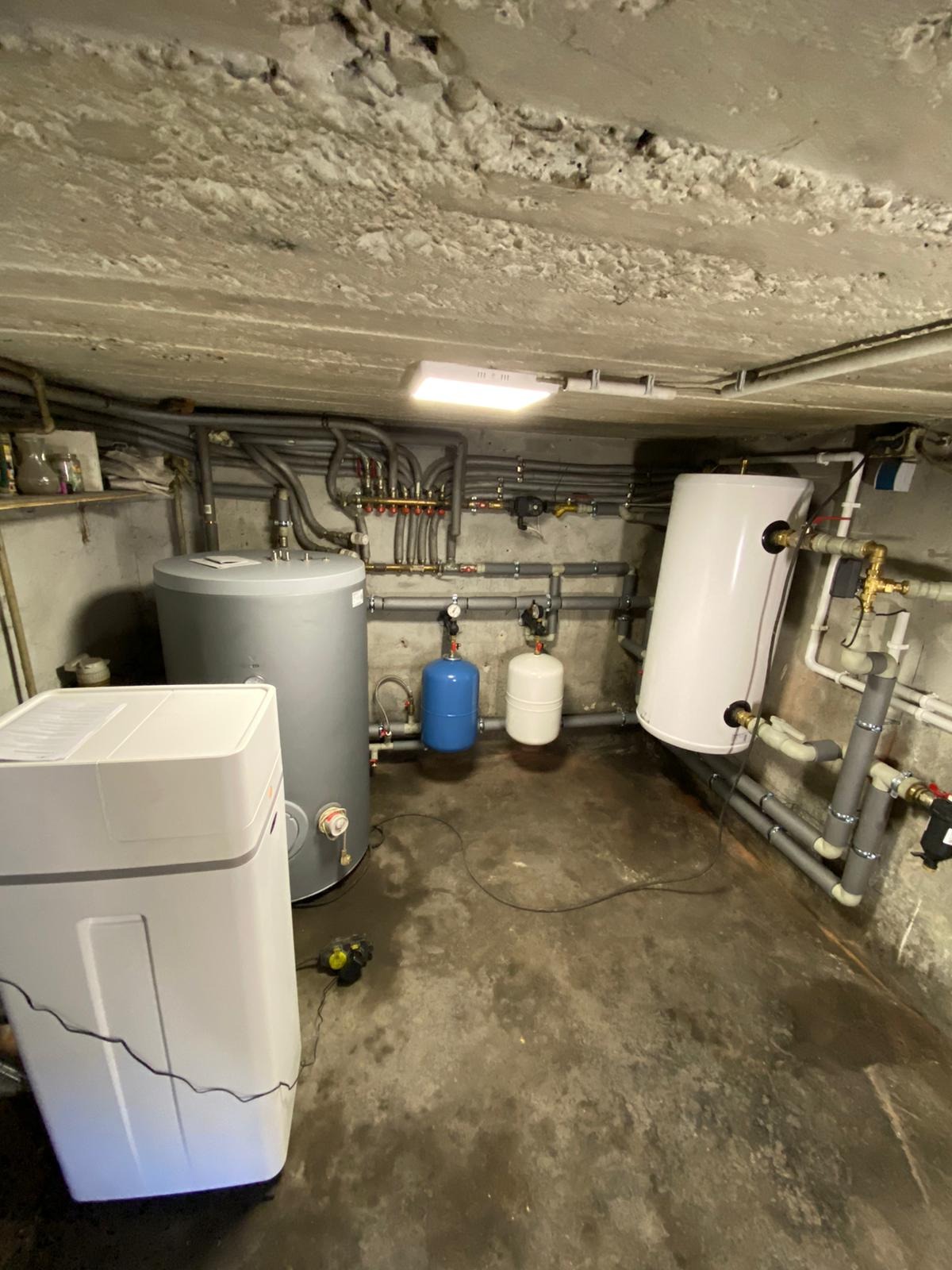 Piwnica z zainstalowanymi zbiornikami na wodę, rozdzielaczem grzewczym i systemem uzdatniania wody. Widoczny surowy betonowy sufit, szare rury instalacyjne i dwa małe zbiorniki wyrównawcze...