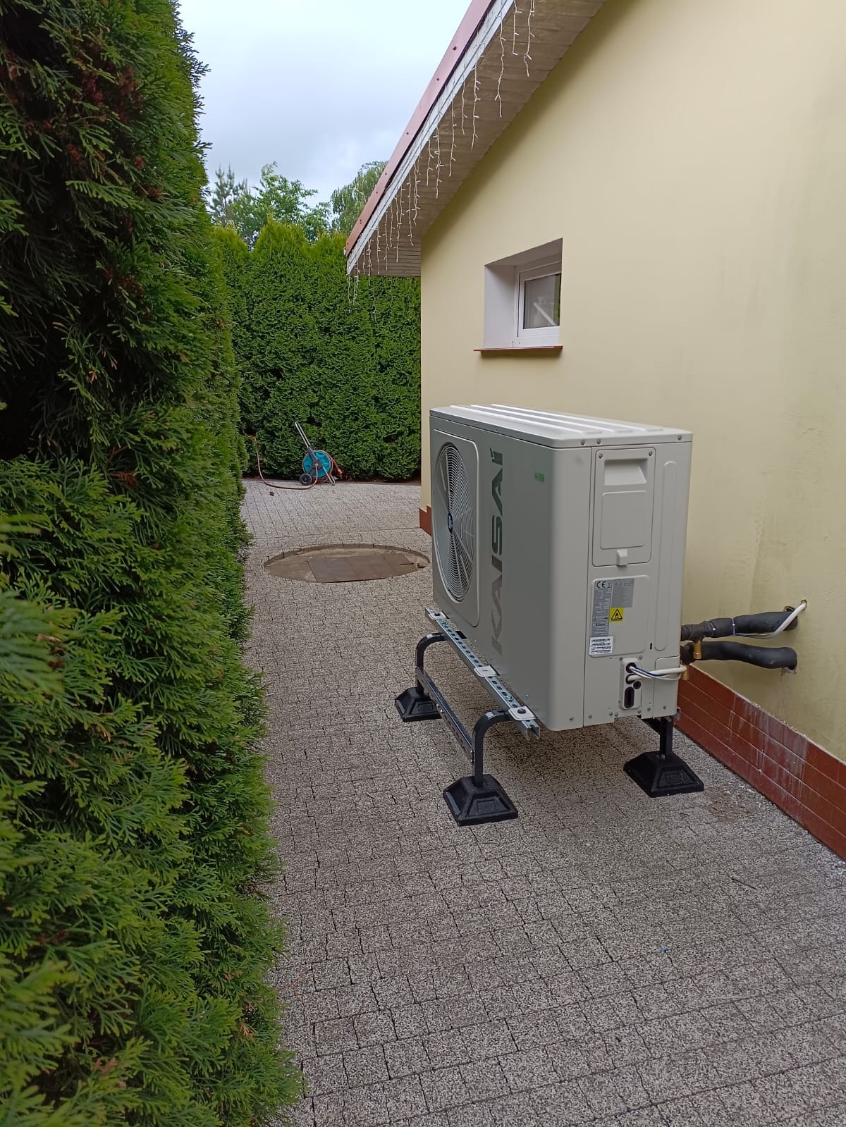Zewnętrzna jednostka pompy ciepła Kaisai zamontowana na metalowym stelażu przy ścianie budynku, widoczne przyłącza rur, w tle tuje i fragment posesji.