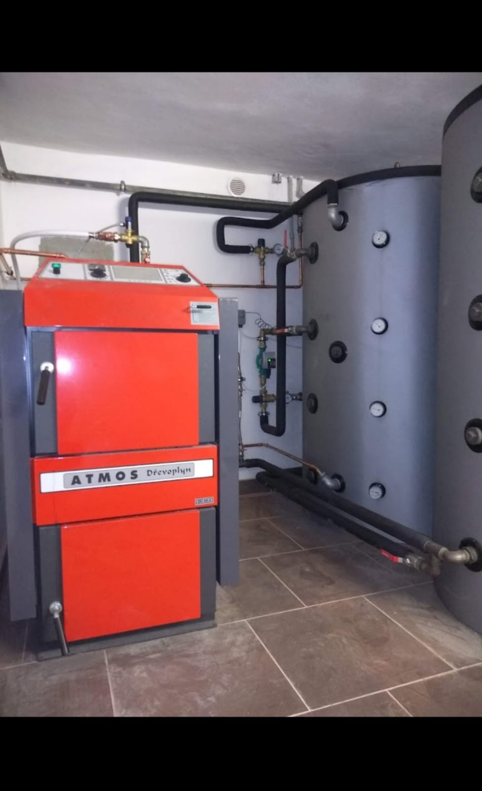 Czerwono-szary piec na paliwo stałe marki Atmos Drevo plyn z podłączonymi rurami i szarym zbiornikiem akumulacyjnym w kotłowni wyłożonej płytkami.
