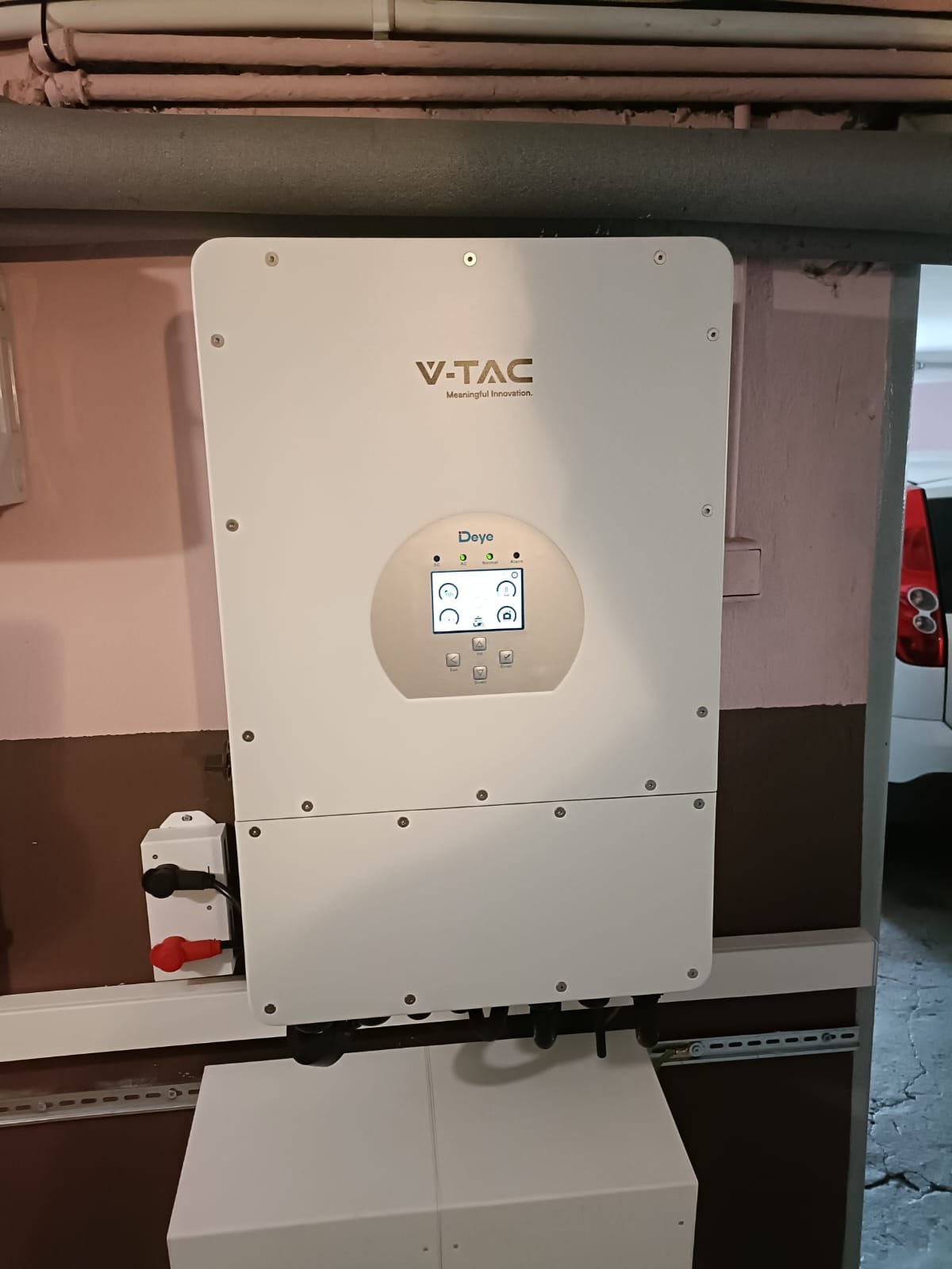 Biały inwerter fotowoltaiczny marki V-TAC zamontowany na ścianie wewnątrz budynku, widoczne przyłącza elektryczne i wyświetlacz pokazujący parametry pracy.