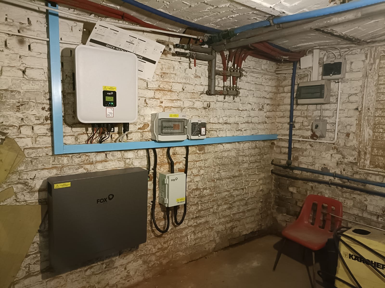 Instalacja magazynu energii Fox ESS i falownika fotowoltaicznego na ścianie z cegły w piwnicy, widoczne rury instalacyjne i skrzynki elektryczne.