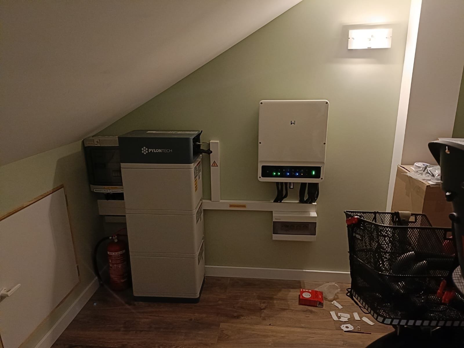 Instalacja magazynu energii Pylontech i falownika w pomieszczeniu ze skośnym sufitem, widoczny także gaśnica i elementy montażowe na podłodze.