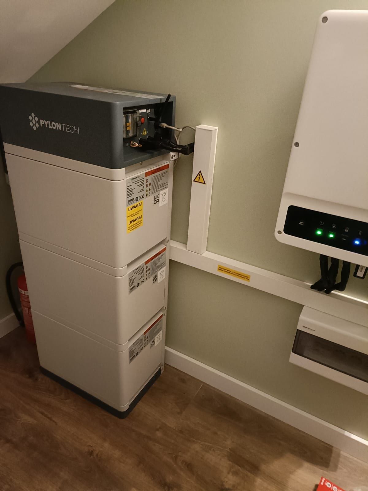 Trzy moduły magazynu energii Pylontech ustawione jeden na drugim, z widocznymi kablami i naklejkami ostrzegawczymi, obok falownik fotowoltaiczny na ścianie w pomieszczeniu z drewnianą podłogą.