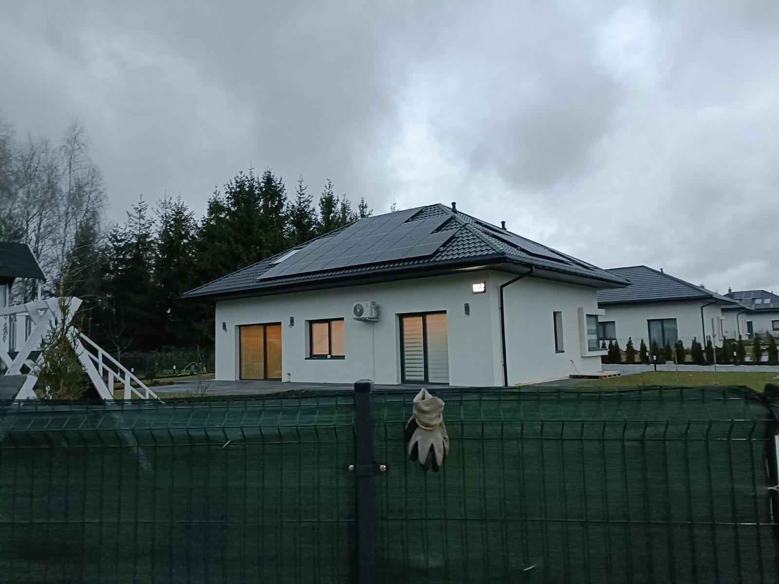 Biały, parterowy dom z panelami słonecznymi na dachu, widziany zza zielonego ogrodzenia z wiszącą roboczą rękawicą. W tle pochmurne niebo i drzewa.