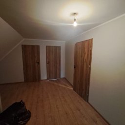 Mirosław Bolda - Trzy drewniane drzwi wewnętrzne z czarnymi klamkami w pokoju na poddaszu z drewnianą podłogą, oświetlonym żarówką.