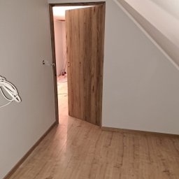 Mirosław Bolda - Otwarta, fornirowana drzwi w nowo wyremontowanym pokoju na poddaszu z panelami podłogowymi, widoczne kable wystające ze ściany, sugerujące niedokończone prace elektryczne.