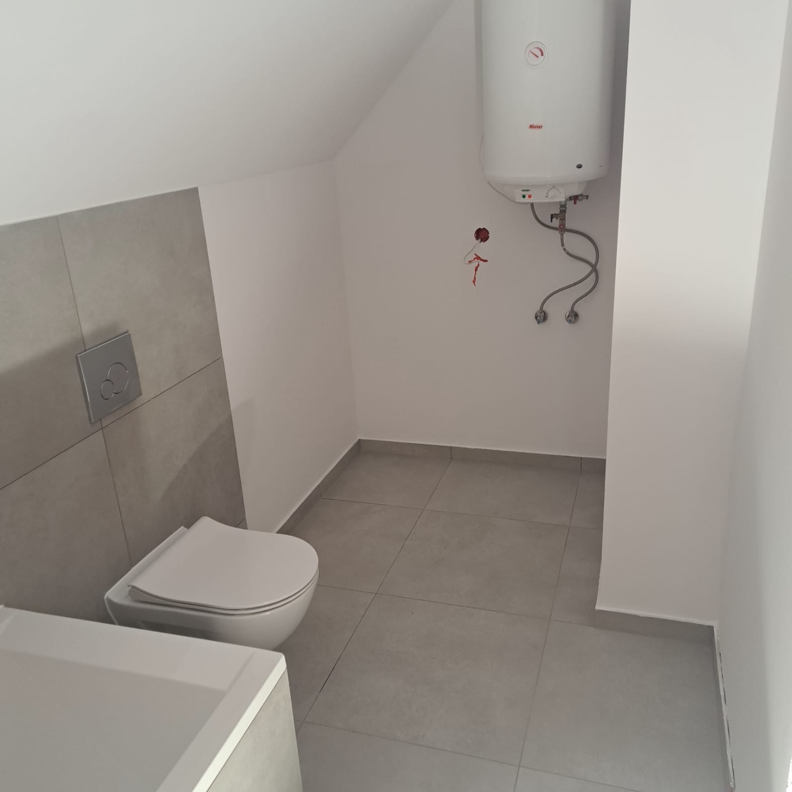 Wnętrze łazienki na poddaszu z szarymi płytkami, podwieszaną toaletą z chromowanym przyciskiem spłukiwania i bojlerem elektrycznym na ścianie, widoczne ślady po instalacji.
